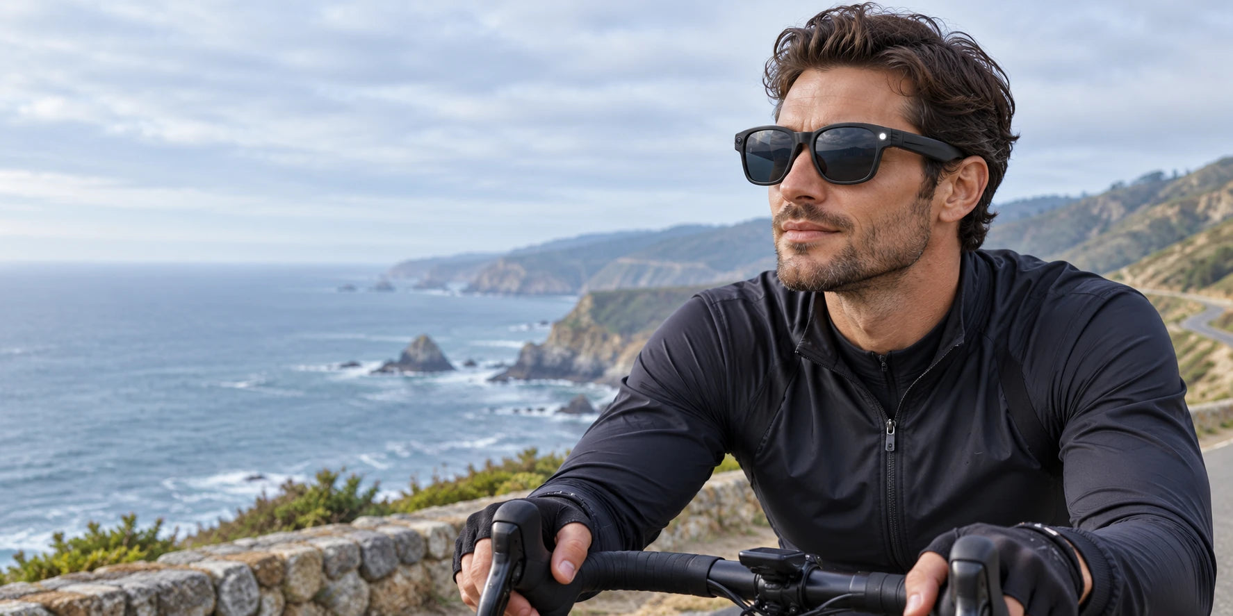 Cyclist wearing black camera sunglasses with both hands on the handlebars at a scenic stop.