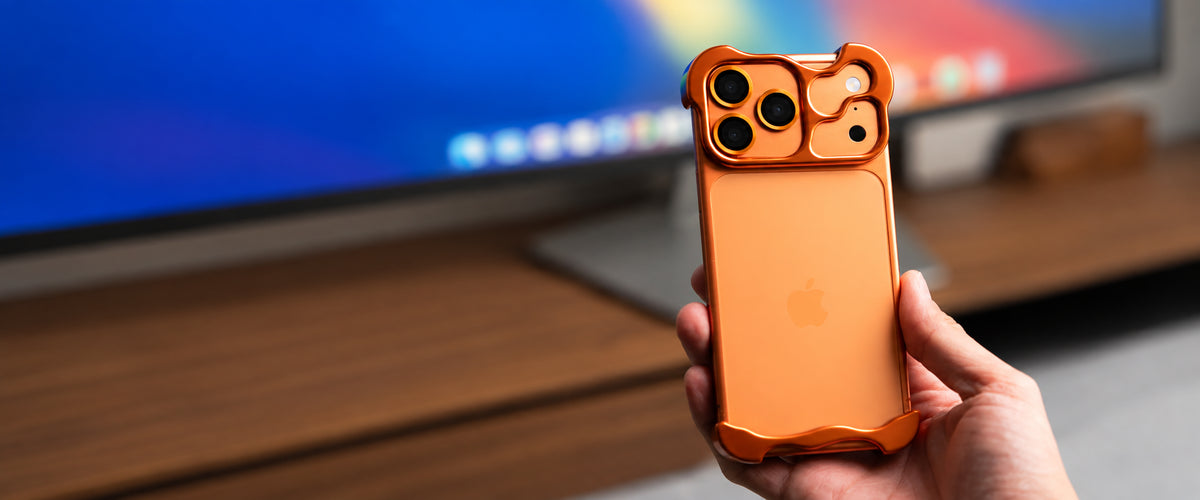 Orange sculpted metal iPhone bumper case held in one hand in front of a desktop monitor.